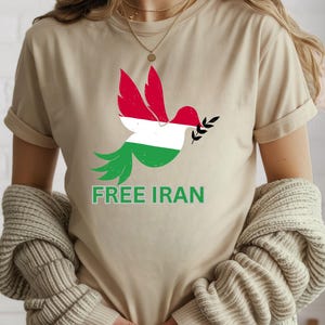 May include: Beige t-shirt featuring a dove graphic with red, white, and green colors, holding a sprig. The text "FREE IRAN" is printed below the dove in green. The shirt is a casual wear item.