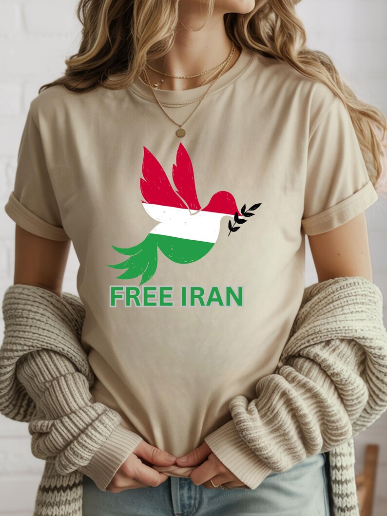 May include: Beige t-shirt featuring a dove graphic with red, white, and green colors, holding a sprig. The text "FREE IRAN" is printed below the dove in green. The shirt is a casual wear item.