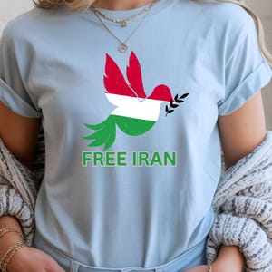 May include: Light blue t-shirt featuring a dove design with the colors of the Iranian flag. The dove holds an olive branch and the words "FREE IRAN" are printed in green below the dove. The shirt has short sleeves.
