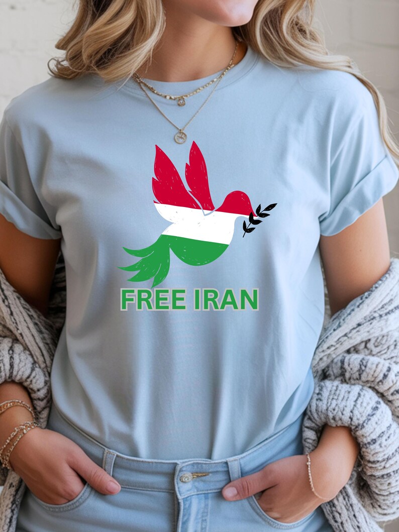 May include: Light blue t-shirt featuring a dove design with the colors of the Iranian flag. The dove holds an olive branch and the words "FREE IRAN" are printed in green below the dove. The shirt has short sleeves.