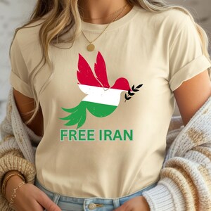 May include: Beige t-shirt featuring a dove design with the colors of the Iranian flag. The dove holds an olive branch and the words "FREE IRAN" are printed in green below the bird. The shirt is made of a soft material.