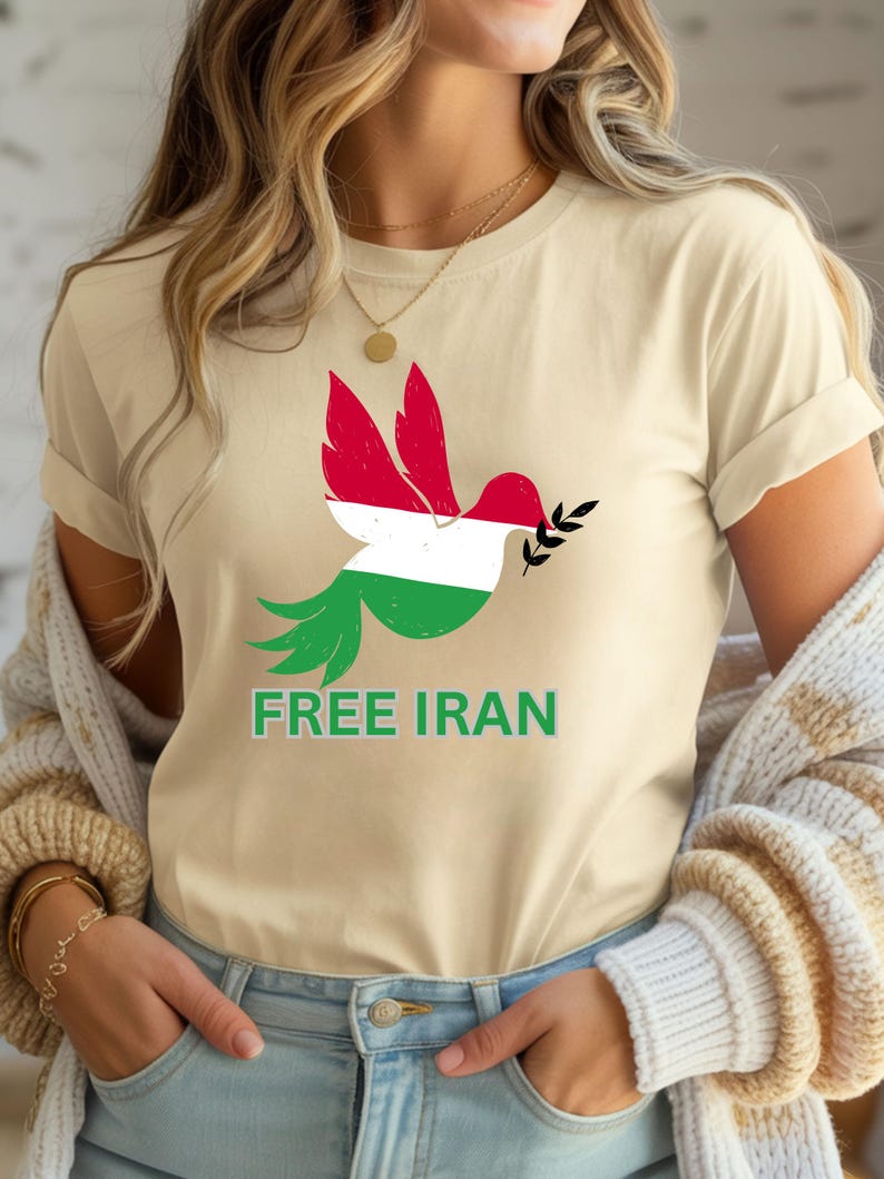 May include: Beige t-shirt featuring a dove design with the colors of the Iranian flag. The dove holds an olive branch and the words "FREE IRAN" are printed in green below the bird. The shirt is made of a soft material.