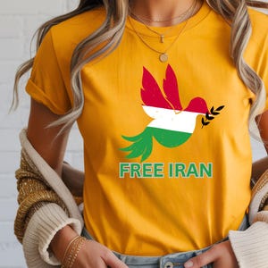 May include: Mustard yellow t-shirt featuring a dove design with the colors of the Iranian flag. The dove holds an olive branch and the words "FREE IRAN" are printed below the bird in green.