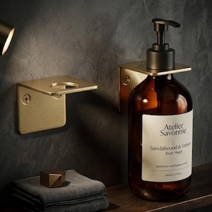 May include: A wall-mounted brushed gold metal holder supporting an amber glass bottle with a black pump dispenser. The label reads Atelier Savonnie Sandalwood & Vetiver Body Wash 100ml / 25 fl oz. An empty matching gold metal wall bracket is adjacent.