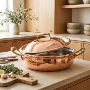 May include: A round, hammered copper cooking pot with a matching lid and two brass side handles. The exterior features a textured, metallic copper finish, while the interior is lined with silver-toned metal. The lid has a central brass handle.