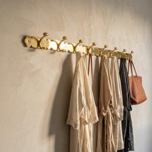 Puede incluir: Un perchero horizontal largo de latón con múltiples ganchos curvos montado en una pared. Colgados de los ganchos hay tres batas ligeras en tonos crema y beige, una bata gris oscuro y un bolso de mano marrón con textura tejida.