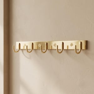 May include: A horizontal brass wall rack featuring five U-shaped hooks. The metal strip has a brushed finish and is secured to the wall with three visible screws positioned between the hooks, providing a functional storage solution.