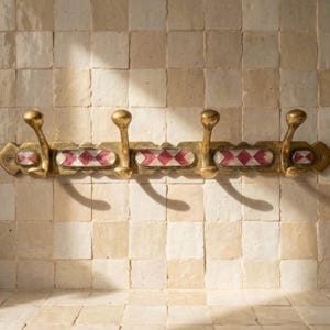 May include: A brass wall-mounted coat rack with four hooks. The rack has oval decorative sections with a red and white diamond pattern. The hooks and rack have a gold finish, adding a touch of elegance.