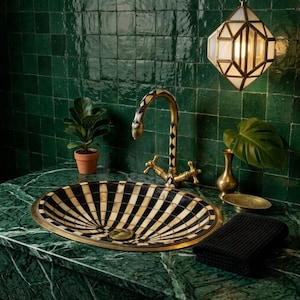 May include: An oval sink basin featuring a black and white radial striped pattern, paired with a matching curved faucet. The setup includes a small potted plant, a brass vase, a black textured towel, and a geometric pendant light fixture.