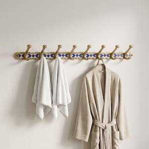 May include: A wall-mounted coat rack with eight brass hooks. The rack has a gold finish with a blue and white geometric pattern. Two white towels and a beige robe hang from the hooks, providing a stylish storage solution for a bathroom or bedroom.