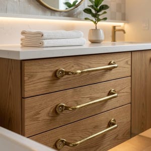 May include: A bathroom vanity cabinet made of light-toned wood with a visible grain, featuring three horizontal drawers. Each drawer is equipped with a long, curved, brushed brass handle secured by two screws. A white countertop sits above.