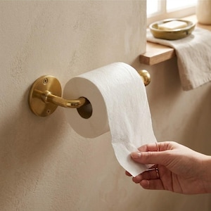 以下が含まれることがあります： 真鍮製の壁掛け式トイレットペーパーホルダー。円形の取り付けプレートと2本のネジが見え、標準的な白いトイレットペーパーのロールを保持しています。金属製のアームが水平に伸びてロールを支える構造になっています。