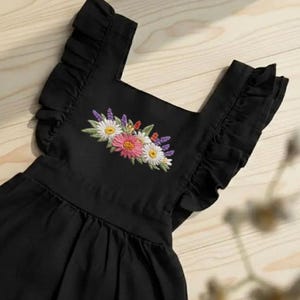 K&ouml;nnte beinhalten: Schwarzes Kleid mit R&uuml;schen&auml;rmeln und eckigem Ausschnitt. Das Kleid hat ein gesticktes Blumendesign in der Mitte, das rosa, wei&szlig;e und gelbe Blumen mit gr&uuml;nen Bl&auml;ttern zeigt. Das Kleid ist aus einem weichen, flie&szlig;enden Stoff gefertigt.