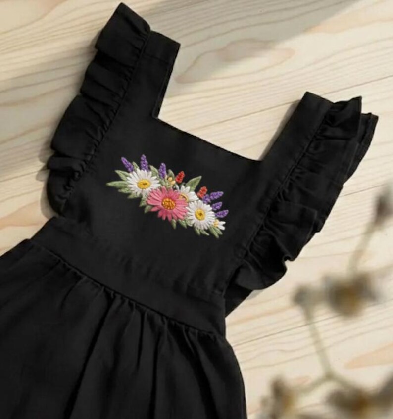 K&ouml;nnte beinhalten: Schwarzes Kleid mit R&uuml;schen&auml;rmeln und eckigem Ausschnitt. Das Kleid hat ein gesticktes Blumendesign in der Mitte, das rosa, wei&szlig;e und gelbe Blumen mit gr&uuml;nen Bl&auml;ttern zeigt. Das Kleid ist aus einem weichen, flie&szlig;enden Stoff gefertigt.