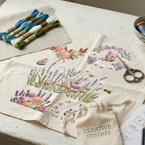 K&ouml;nnte beinhalten: Eine Bastelszene mit Stickzubeh&ouml;r. Ein floral gesticktes Design auf Leinen ist umgeben von Stickgarn, einer kleinen Schale mit Faden, einem Nadelkissen, einer Schere und einem kleinen Beutel mit dem Text "CREATIVE STITCHES".