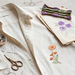 K&ouml;nnte beinhalten: Ein cremefarbenes Hemd mit Kn&ouml;pfen und einem gestickten orangefarbenen Blumendesign. Das Bild zeigt auch Stickgarn, Nadeln, eine Schere und eine Blumenskizze, was auf ein Bastelthema hindeutet.