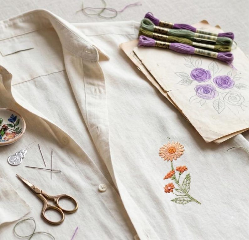 K&ouml;nnte beinhalten: Ein cremefarbenes Hemd mit Kn&ouml;pfen und einem gestickten orangefarbenen Blumendesign. Das Bild zeigt auch Stickgarn, Nadeln, eine Schere und eine Blumenskizze, was auf ein Bastelthema hindeutet.