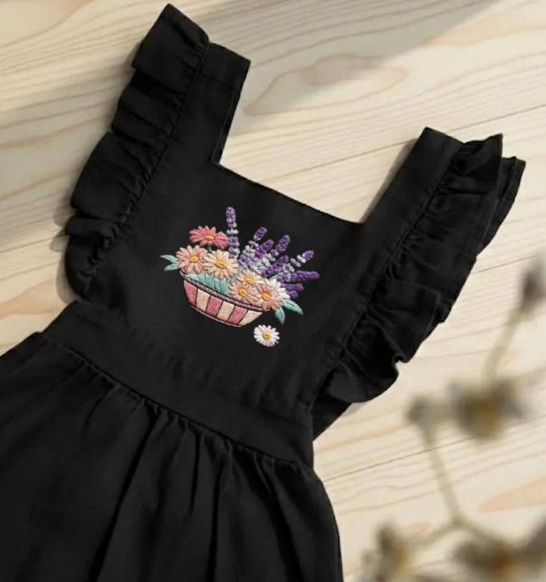K&ouml;nnte beinhalten: Schwarzes Kleid mit R&uuml;schen&auml;rmeln und eckigem Ausschnitt. Das Kleid hat ein gesticktes Blumendesign in einem Korb, mit rosa, gelben und lila Blumen. Unter dem Korb ist eine kleine wei&szlig;e G&auml;nsebl&uuml;mchen gestickt.