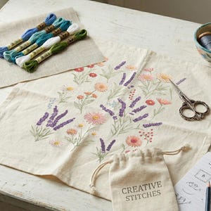 K&ouml;nnte beinhalten: Cremefarbener Stoff mit gesticktem Blumendesign, darunter Lavendel und G&auml;nsebl&uuml;mchen. Stickgarn in Blau-, Gr&uuml;n- und Wei&szlig;t&ouml;nen, eine kleine Schale mit Garn, eine Schere und ein Nadelkissen deuten auf ein Handarbeitsprojekt hin.