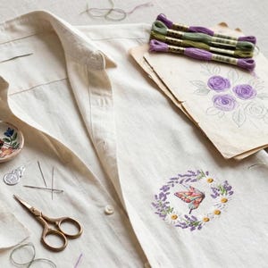 K&ouml;nnte beinhalten: Ein cremefarbenes Leinenhemd mit einem gestickten Schmetterlings- und Blumendesign. Das Bild zeigt Stickgarn, Nadeln, eine Schere und eine Blumenskizze. Der Schmetterling ist von einem Kranz aus Lavendel und G&auml;nsebl&uuml;mchen umgeben.