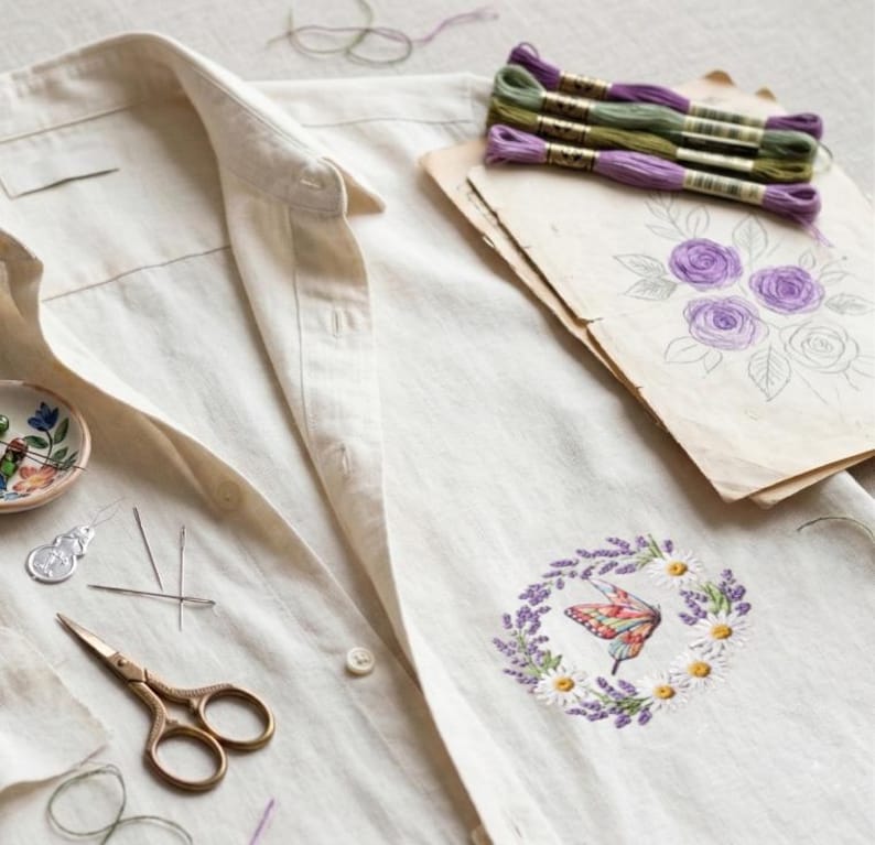 K&ouml;nnte beinhalten: Ein cremefarbenes Leinenhemd mit einem gestickten Schmetterlings- und Blumendesign. Das Bild zeigt Stickgarn, Nadeln, eine Schere und eine Blumenskizze. Der Schmetterling ist von einem Kranz aus Lavendel und G&auml;nsebl&uuml;mchen umgeben.