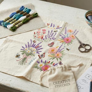 K&ouml;nnte beinhalten: Ein florales Stickprojekt mit einem cremefarbenen Stoff mit einem farbenfrohen Blumenstrau&szlig;-Design. Enth&auml;lt Stickgarn, eine kleine Schale mit Faden, eine Schere, ein Nadelkissen und einen kleinen Beutel mit dem Text "CREATIVE STITCHES".