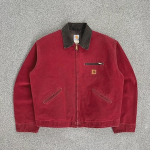 Puede incluir: Chaqueta Carhartt roja con cuello marrón oscuro y cierre de cremallera. La chaqueta tiene dos bolsillos delanteros y un pequeño bolsillo con cremallera en el pecho. Un parche con el logotipo de Carhartt es visible en el pecho.