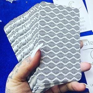 May include: A hand holds a deck of cards featuring a black and white wavy design. The cards have rounded edges. Other cards with French text are visible in the background. The background is dark blue.