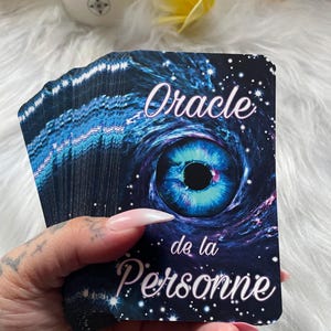 Peut inclure: Un jeu de cartes oracle avec un design d'œil cosmique bleu et violet. Les cartes sont tenues dans une main avec de longs ongles manucurés. Le texte "Oracle de la Personne" est visible sur la carte.
