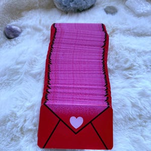 May include: A tall, vertical stack of rectangular cards with rounded corners. The top card features a bright red envelope design with a central light pink heart icon. The edges of the cards in the stack are a vibrant pink, creating a distinct color contrast.