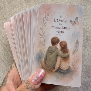 May include: A deck of oracle cards featuring a watercolor illustration of two figures sitting together. The top card displays the text L'Oracle des traumatismes vécus, surrounded by a delicate frame of leaves, a butterfly, and soft pastel washes of pink and blue.