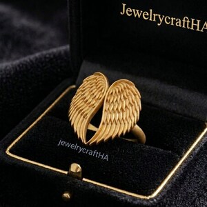 May include: A gold-toned ring featuring two symmetrical angel wings meeting at the center, displayed inside a black velvet jewelry box with the text JewelrycraftHA printed in gold on the interior lid and on the base of the ring setting.