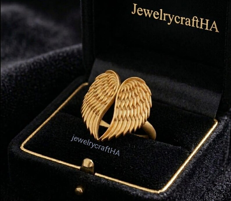 May include: A gold-toned ring featuring two symmetrical angel wings meeting at the center, displayed inside a black velvet jewelry box with the text JewelrycraftHA printed in gold on the interior lid and on the base of the ring setting.
