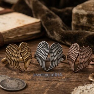 May include: Three rings featuring a pair of angel wings design, arranged in a row. The rings are finished in antique brass, silver, and copper tones. The text jewelrycraftHA is visible in the center of the image below the silver ring.
