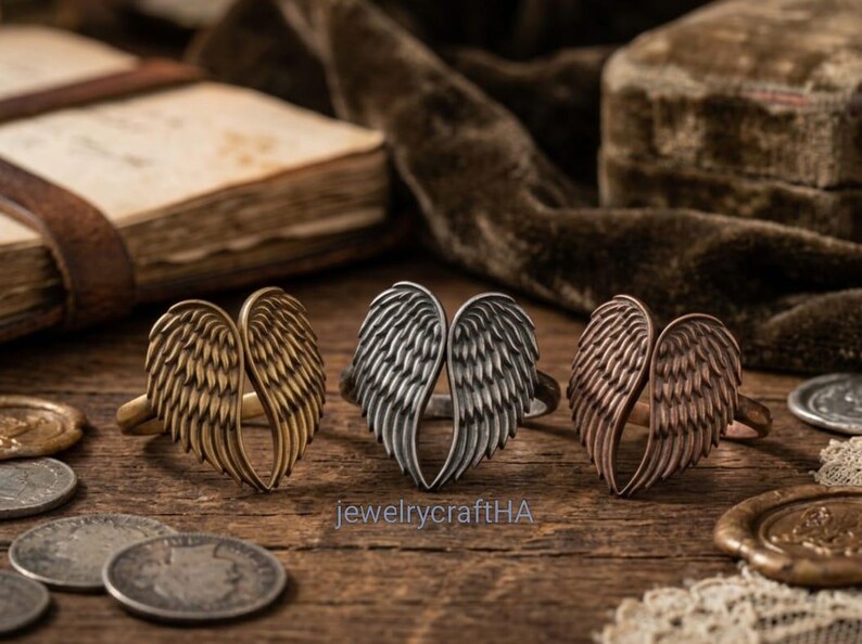 May include: Three rings featuring a pair of angel wings design, arranged in a row. The rings are finished in antique brass, silver, and copper tones. The text jewelrycraftHA is visible in the center of the image below the silver ring.