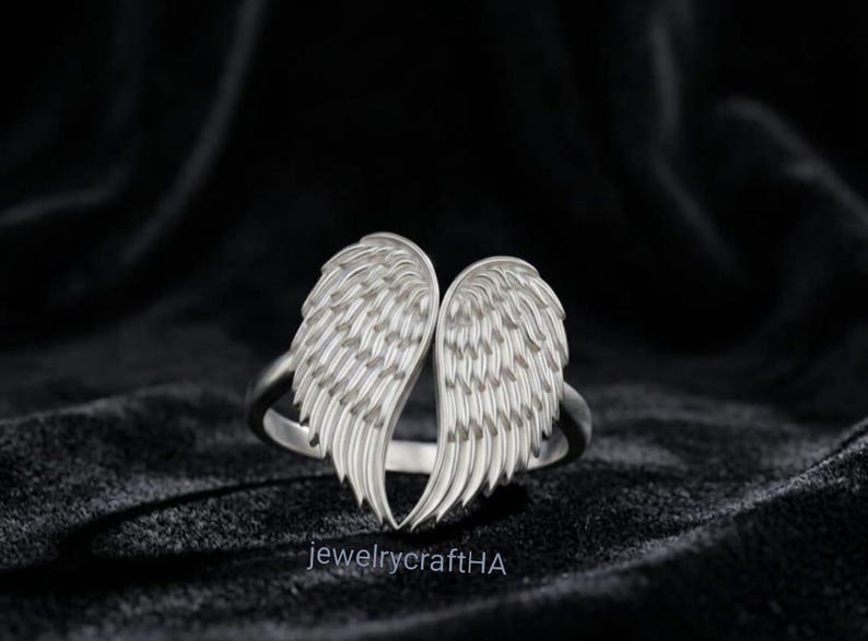 May include: A silver-toned ring featuring two symmetrical, detailed angel wings that meet at the center. The wings have textured, layered feather patterns. The text jewelrycraftHA is printed in small, white letters at the bottom of the image.