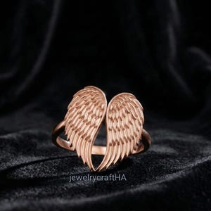 May include: A rose gold ring featuring two symmetrical angel wings that meet at the center to form a heart shape. The wings have detailed, engraved feather textures throughout. The text jewelrycraftHA is printed in small white letters at the bottom.