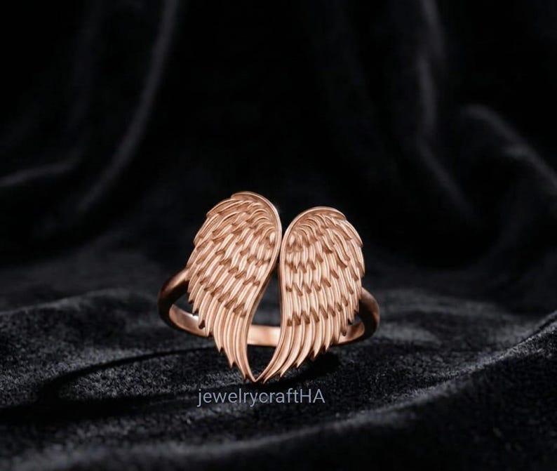 May include: A rose gold ring featuring two symmetrical angel wings that meet at the center to form a heart shape. The wings have detailed, engraved feather textures throughout. The text jewelrycraftHA is printed in small white letters at the bottom.
