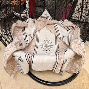 May include: A beige and brown patterned textile draped over a black chair. The textile features vertical brown stripes with black and red accents, and a central geometric design. The fabric appears to be woven.