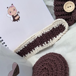 Könnte beinhalten: Handgefertigter Häkel-Lesezeichen mit dunkelbrauner Mitte und cremefarbenen Rändern, neben einem passenden Untersetzer und Stirnband. Eine weiße Tasse mit Herzdesign und ein Notizbuch mit Bienenillustration sind ebenfalls zu sehen.