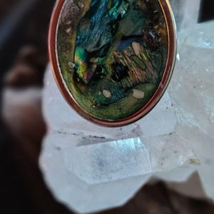 Może przedstawiać: Owalny pierścionek ze srebrną oprawą i kolorową inkrustacją z muszli abalonu. Muszla prezentuje opalizujące odcienie niebieskiego, zielonego i złota. Pierścionek umieszczony jest na tle przezroczystych kryształów kwarcu, tworząc uderzający kontrast.