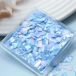 May include: A clear, square container filled with iridescent, light blue flakes. The flakes have a shimmering, pearlescent quality, reflecting hints of pink and purple. The container sits on a light blue surface.
