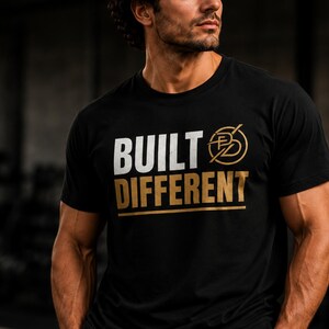 Könnte beinhalten: Ein schwarzes Kurzarm-T-Shirt mit dem Text BUILT in Weiß und DIFFERENT in goldener Schrift. Rechts neben dem Wort BUILT befindet sich ein kreisförmiges Logo mit den Buchstaben BD in Gold. Unter dem Text befindet sich eine goldene Linie.