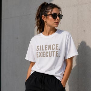 May include: A white short-sleeved t-shirt featuring the text SILENCE. EXECUTE. printed in a neutral beige color across the chest. The garment has a classic crew neckline and a straight hem, constructed from a smooth, opaque cotton fabric.