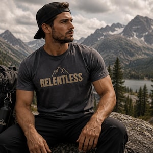 Peut inclure: Un t-shirt gris anthracite avec le mot "RELENTLESS" en beige, orné d'un motif de montagne. Un sac à dos et une gourde noirs sont visibles. La scène se déroule sur fond de montagnes et de lac.