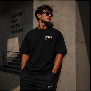 May include: A black short-sleeved t-shirt featuring the text NEVER. SETTLE. printed in white and gold on the chest. The shirt is paired with black shorts that have the word LEGGOD printed in white on the lower left leg area.