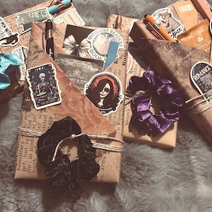 May include: Several gift boxes wrapped in brown paper with stickers and scrunchies. The boxes are tied with twine and adorned with stickers featuring illustrations and text. Scrunchies in blue, purple, and orange are visible.