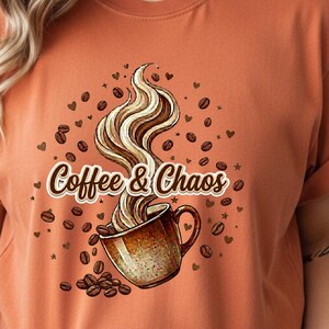 May include: A burnt orange short-sleeve t-shirt featuring a graphic of a coffee mug with swirling steam. The text Coffee & Chaos is printed across the center, surrounded by scattered coffee beans and small heart shapes in a brown color palette.