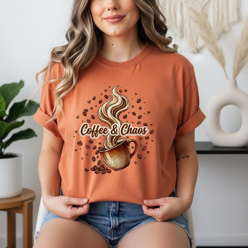 May include: A burnt orange short-sleeve t-shirt featuring a graphic of a coffee mug with swirling steam. The text Coffee & Chaos is printed across the center, surrounded by scattered coffee beans and small heart shapes in a brown color palette.