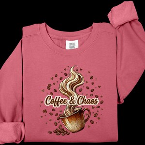 May include: A dusty rose long-sleeve sweatshirt featuring a graphic of a brown coffee mug with swirling steam, surrounded by scattered coffee beans and small hearts. The text Coffee & Chaos is printed in a brown script font across the center.